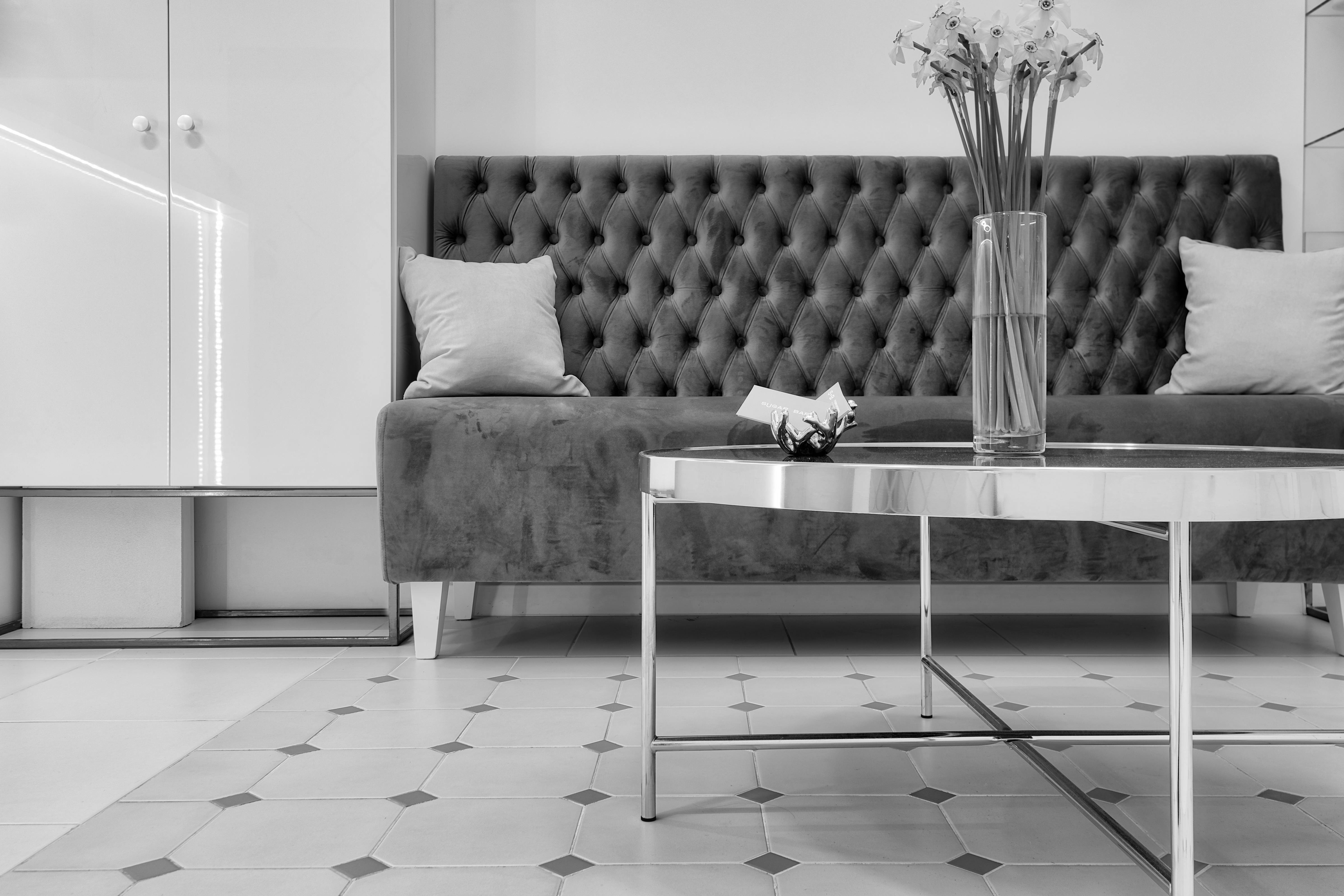 A tufted velvet settee and polished chrome-and-glass coffee table in a bright, tiled room, in black and white.